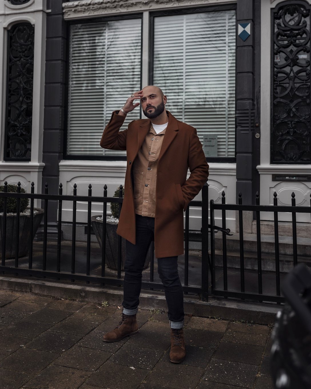 Man In Brown Coat And Denim Jeans Standing Near Black Metal Gate